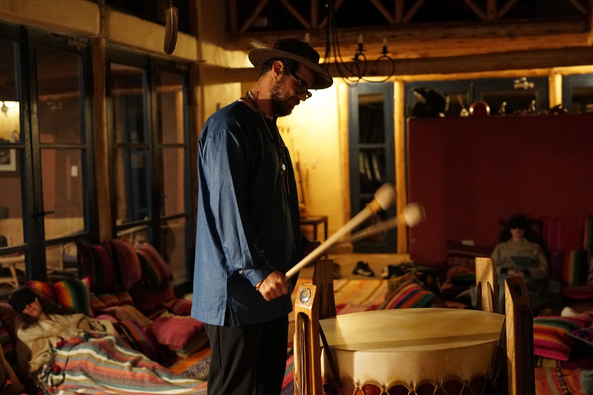 A facilitator plays a large ceremonial drum while the group rests in the shala at Melissa Wasi